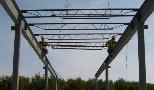 Two construction workers with safety vest and hard hats atop two parallel steel beams.