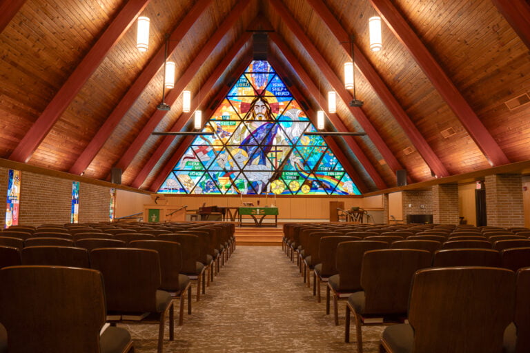 Interior of large A-framed church sanctuary with large ceiling beams and wood paneling on the ceiling, with chairs underneath. Pulpit stage area to the back with a large stained glass religious window in the background.