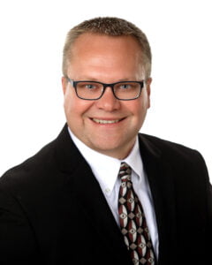 Middle-aged white man with short blonde spikey hair, black rectangular-framed glasses, blue eyes and a smile wearing a black suite and white button-up shirt and geometric tie.