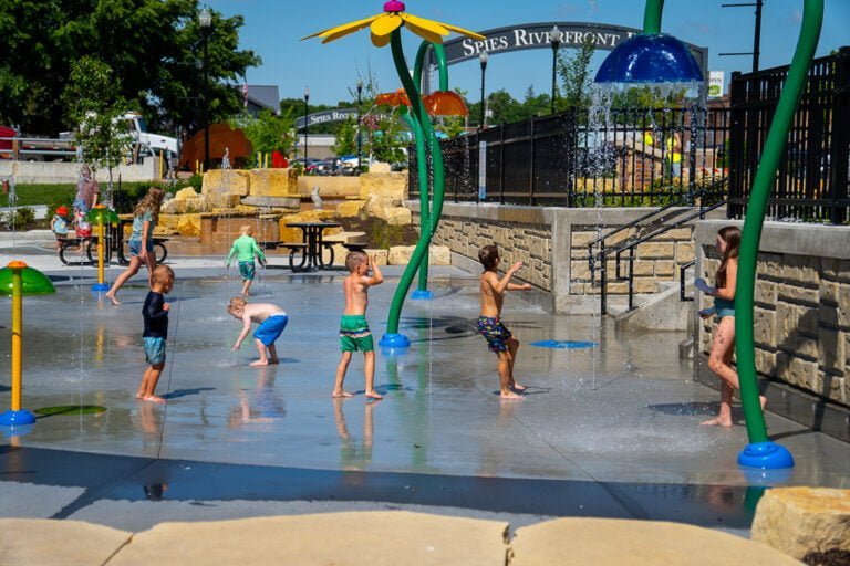 Brightly colored splash pad sprayers and spouts with children in swimsuits running underneath with the "Spies Riverfront Park" sign in the background.