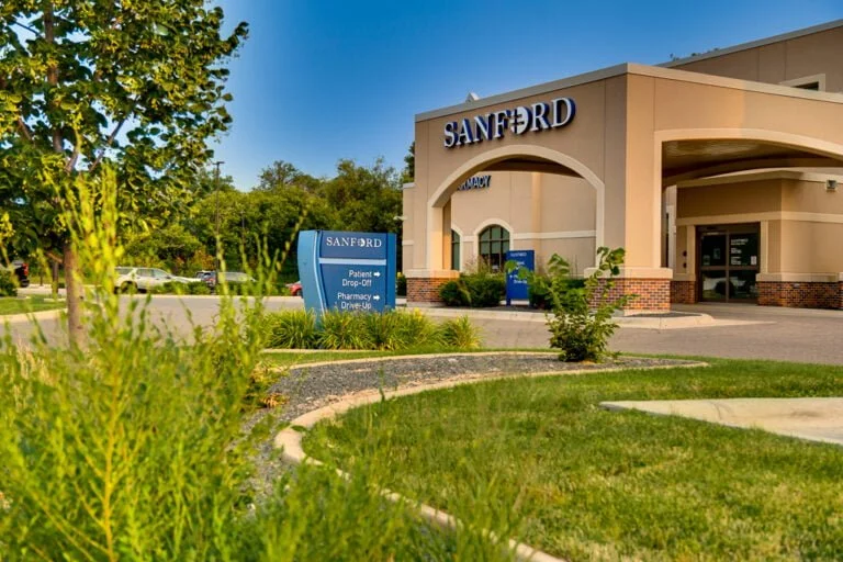 Exterior of beige-colored stucco building with porte-cochère with the word "Sanford" in white letters atop. Blue sky in background and green grass in front with shrubs, trees, and grass for landscaping.