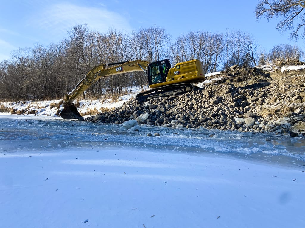 Excavator moving rocks alongside an ice-coated riverbank to cover stream barbs and prevent erosion.