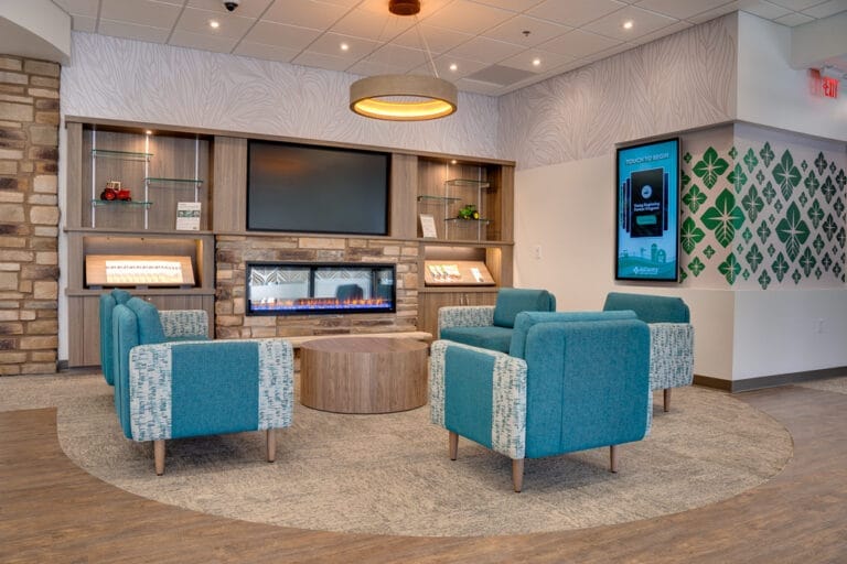 Front waiting area of AgCountry Farm Credit Services in Lamoure, ND, with brick facade electric fireplace and cabinetry flanking each side, aqua blue seating chairs and coffee tables in front, and electronic interactive display monitor to the side.